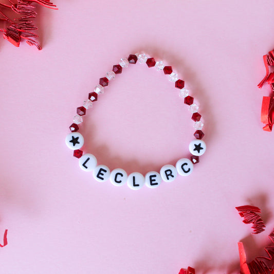 A beaded bracelet spelling 'LECLERC' with white letter beads and red faceted beads, arranged in a circle on a pink background with red tinsel accents.